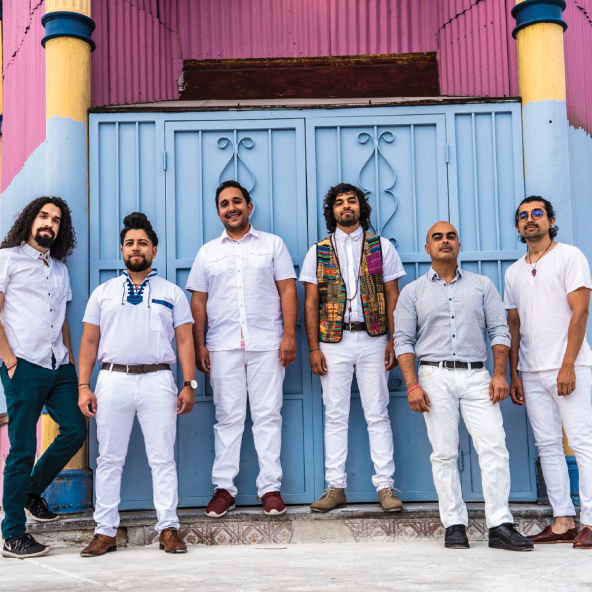 6 musicians in white clothes standing against a blue gate.